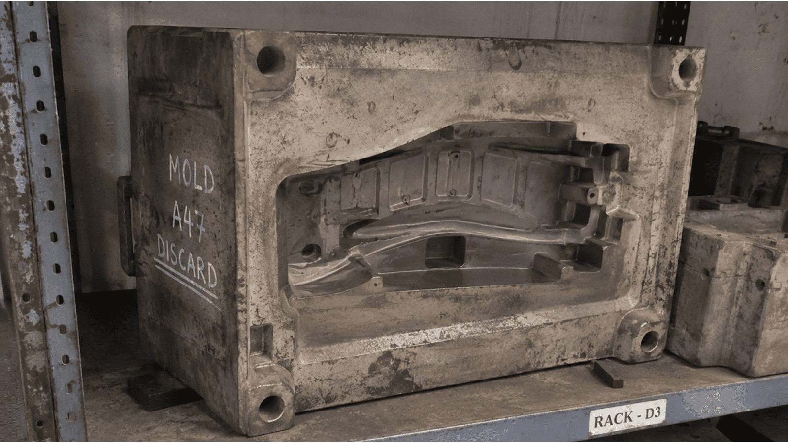 13-Part-5-Mould-Decommissioning-1 - Efficient-Innovations Dusty industrial metal mold labeled 'MOLD A47 DISCARD' on a shelf, with a 'RACK - D3' tag underneath on the rack edge.