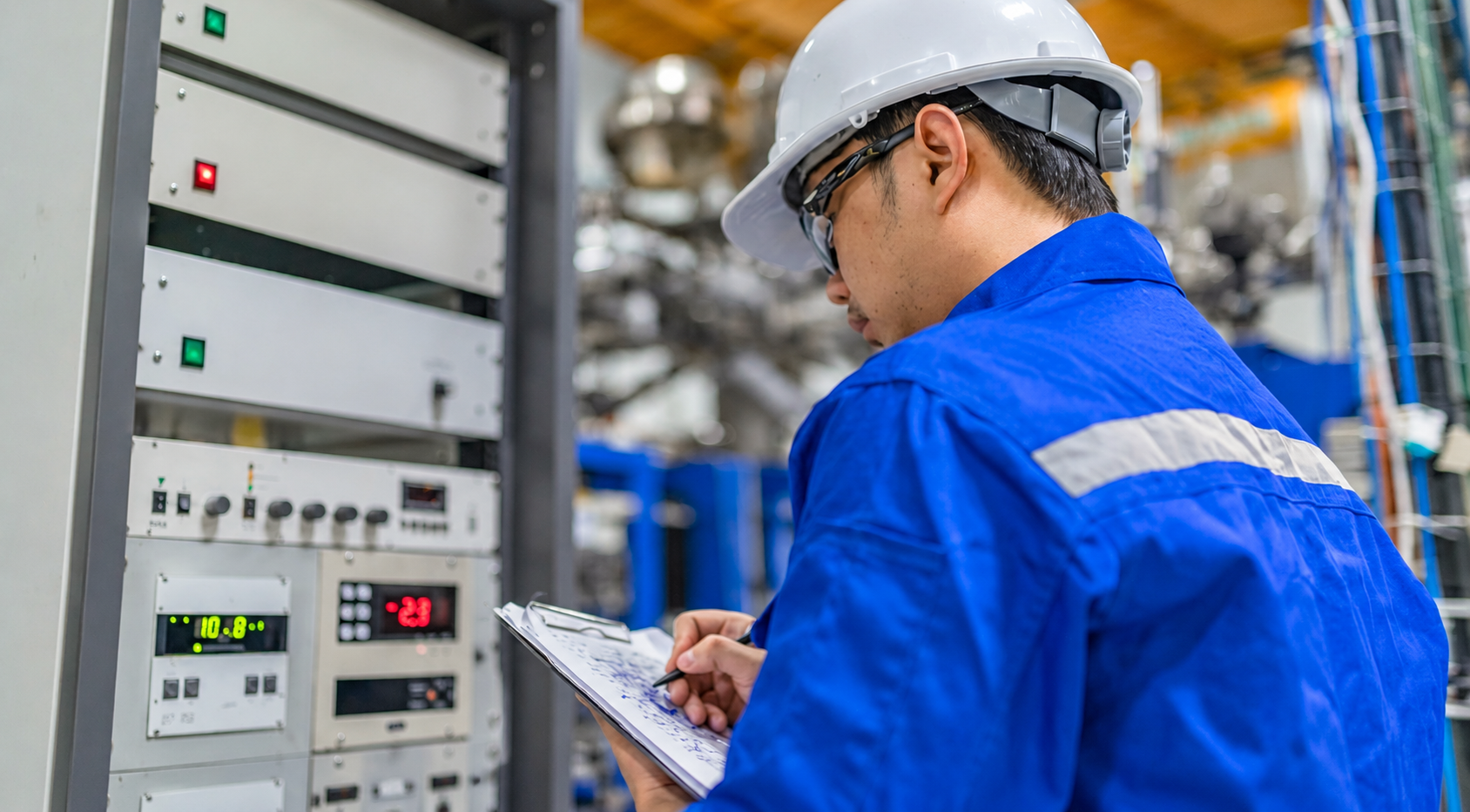Engineer in a white hard hat and blue coverall takes notes on a clipboard beside an industrial control panel.