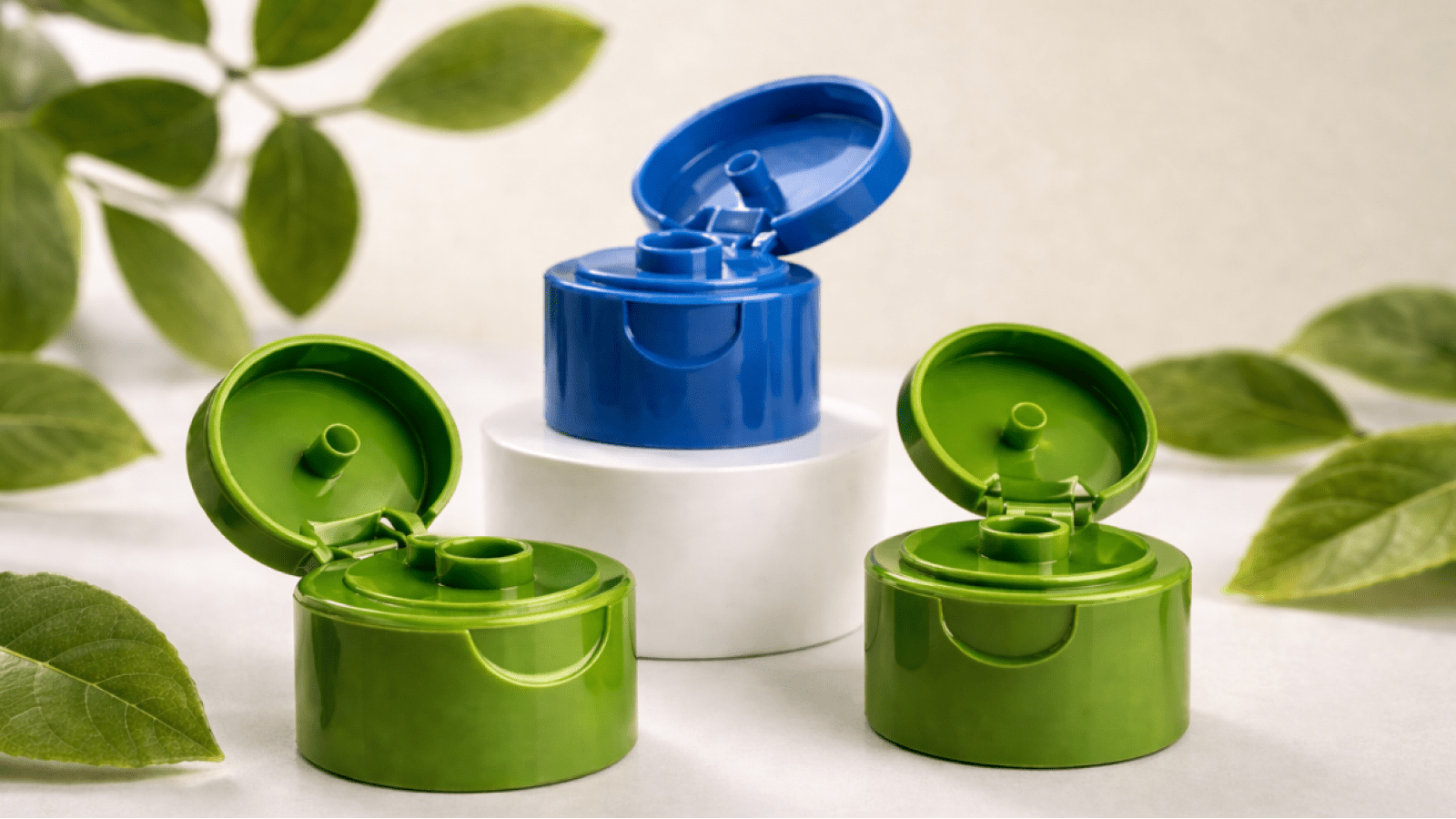 Three plastic bottle caps (two green, one blue) arranged on a white display, blue cap open while green caps are closed, with green leaves in the background.