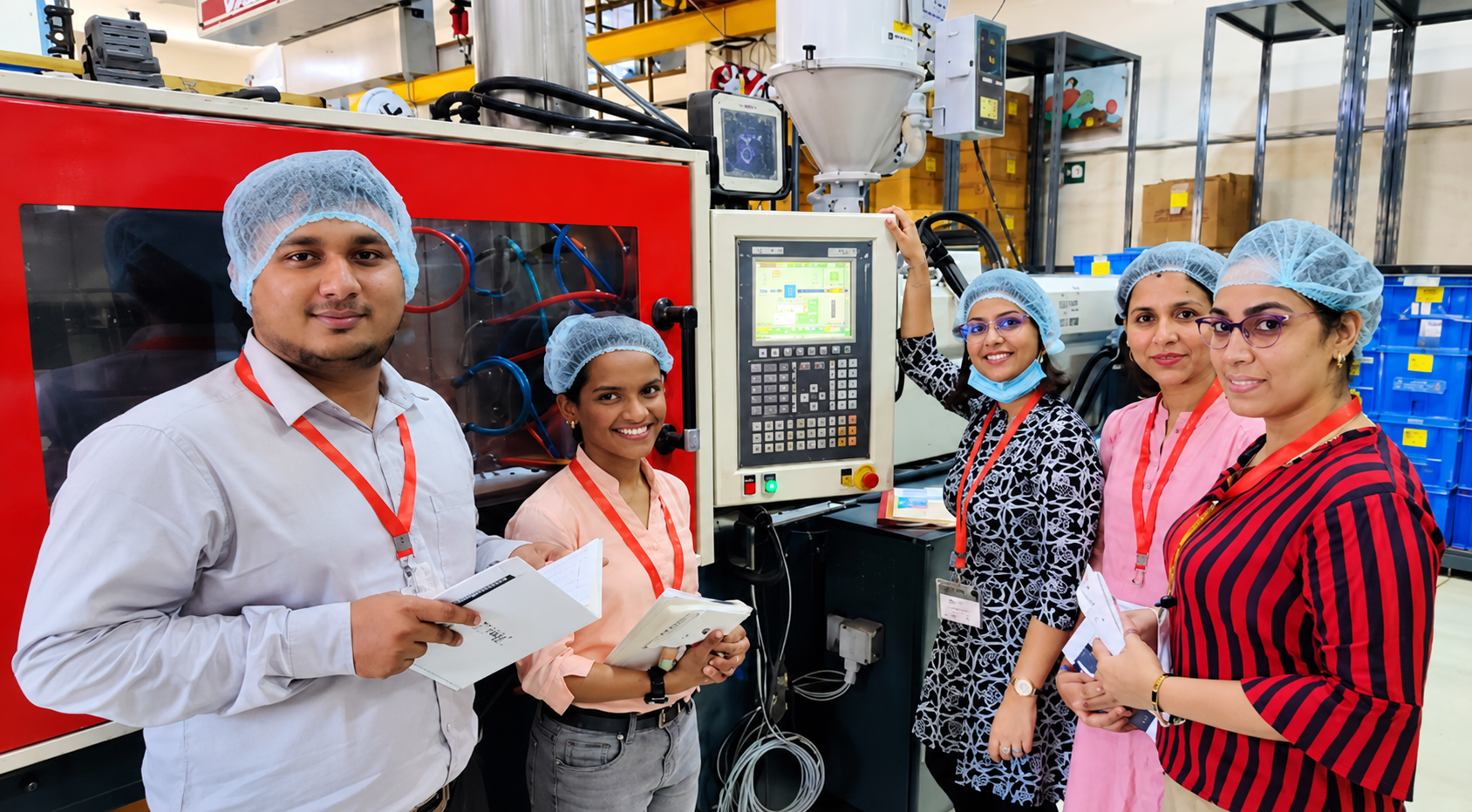 The Research Road Less Travelled for Injection Moulding IM Process Development - Efficient-Innovations Five factory workers wearing hair nets and red lanyards stand beside a large control-panel machine in a manufacturing plant, smiling at the camera.