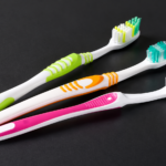 Three colorful plastic toothbrushes with white handles lying on a dark surface, green, orange, and pink accents visible.