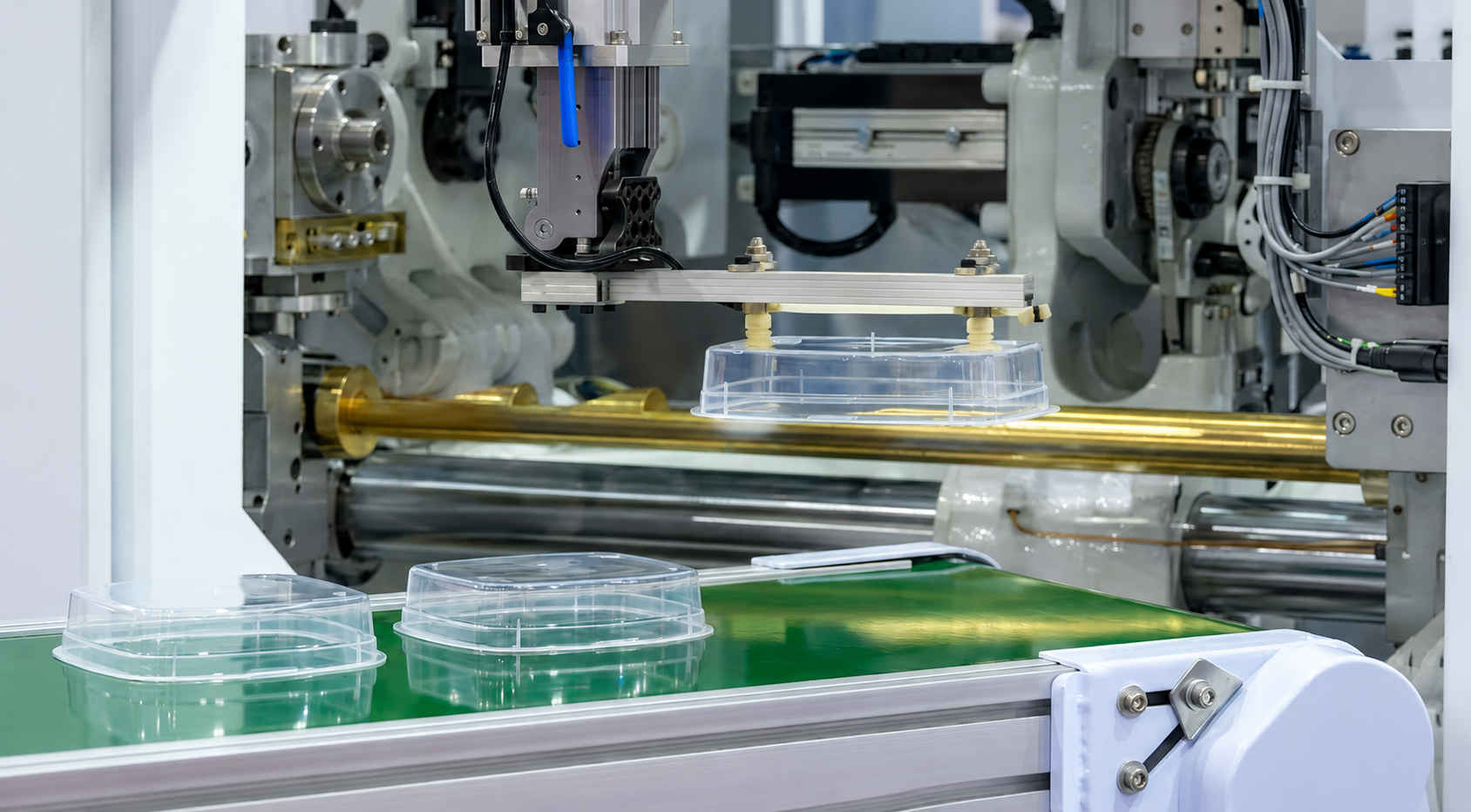 Robotic arm placing clear plastic clamshell lids onto circular containers on a packaging line with a green conveyor belt.