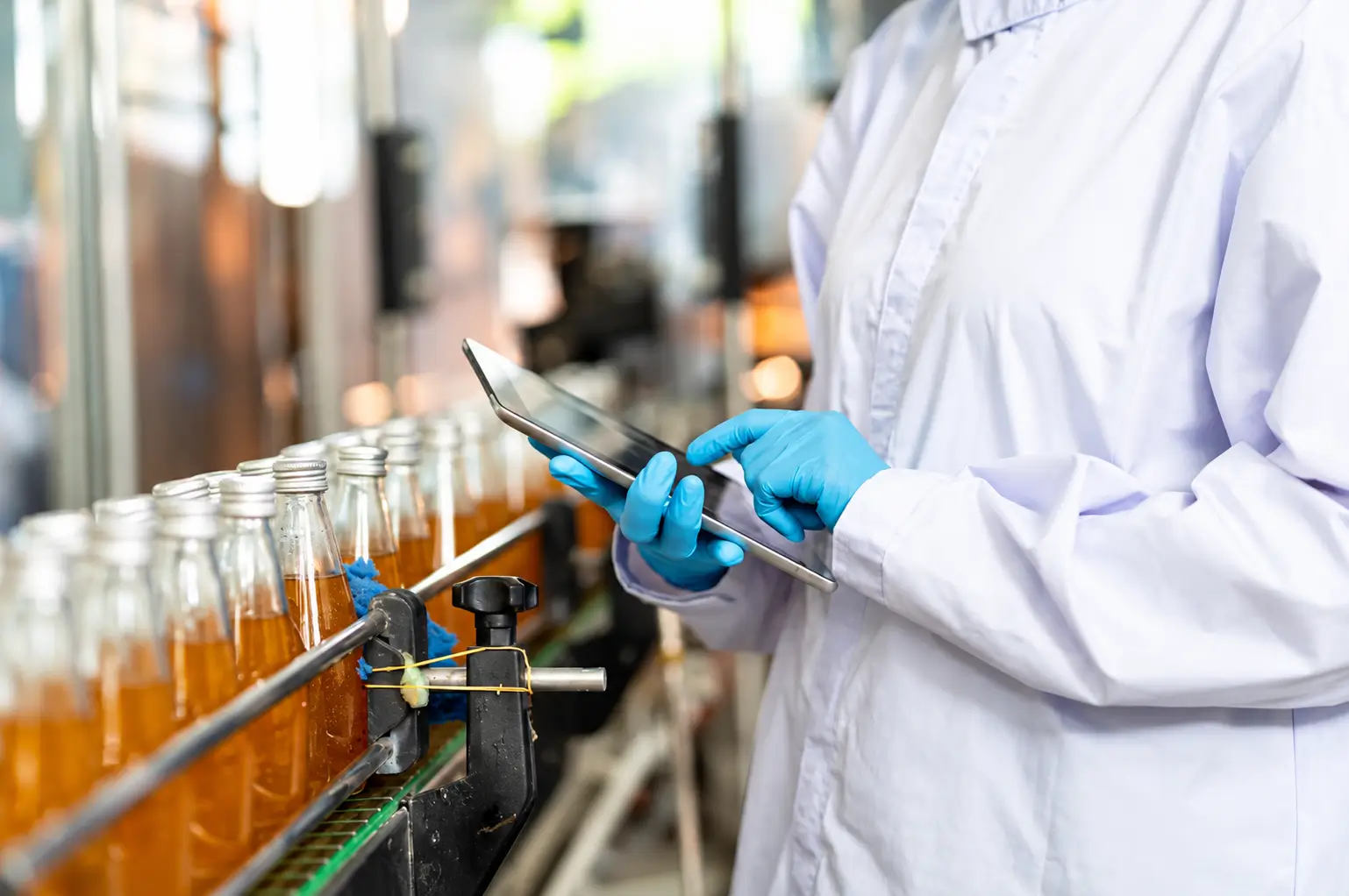 Lab technician in a white coat using a tablet beside a row of orange beverage bottles on a conveyor.
