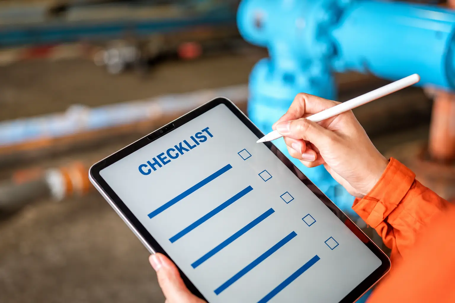 Person using a stylus to fill out a maintenance checklist on a tablet in an industrial setting with blue machinery in the background.