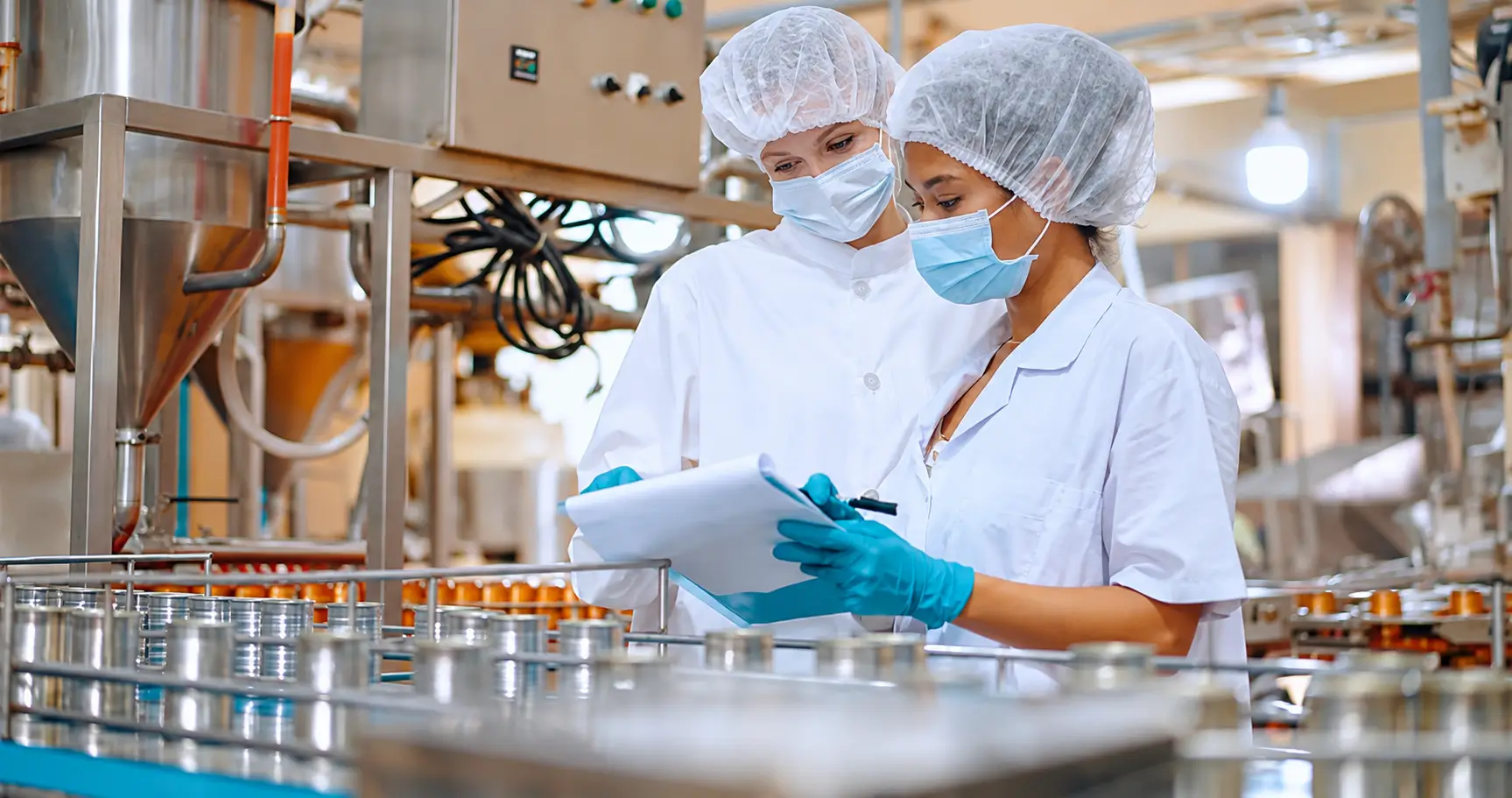 Two factory workers in masks, hair nets, and blue gloves examine documents at a production line.