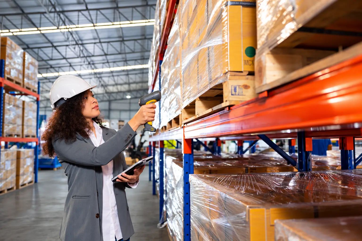 Warehouse worker in a hard hat scans wrapped pallets with a handheld scanner, checking inventory.